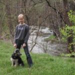 Eine Person steht im Gras neben einem Hund, im Hintergrund sind ein kleiner Fluss und Bäume zu sehen.