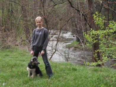 Eine Person steht im Gras neben einem Hund, im Hintergrund sind ein kleiner Fluss und Bäume zu sehen.