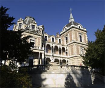 Großes historisches Herrenhaus mit verschnörkelter Architektur, Ziegel- und Steinfassade, zahlreichen Fenstern, Balkonen, Türmen und einem Spitzdach, umgeben von Bäumen unter einem klaren blauen Himmel.