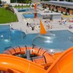 Blick auf ein Freibad mit mehreren Schwimmbecken, Lounge-Bereichen und leuchtend orangefarbenen Wasserrutschen. Menschen nehmen ein Sonnenbad und schwimmen.