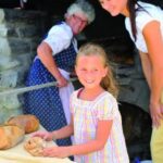 Eine ältere Frau backt Brot in einem Steinofen, während ein lächelndes Mädchen und eine Frau in der Nähe stehen und Brote auf einem Tisch vor sich haben.