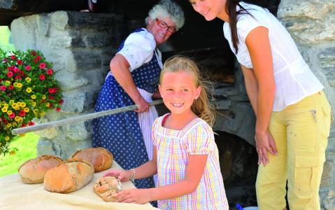 Eine ältere Frau backt Brot in einem Steinofen, während ein lächelndes Mädchen und eine Frau in der Nähe stehen und Brote auf einem Tisch vor sich haben.