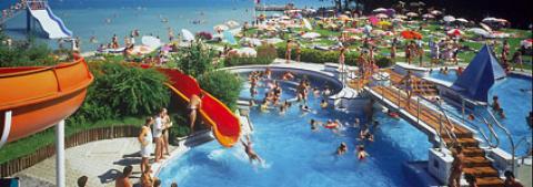 Menschen schwimmen und spielen auf Wasserrutschen in einem überfüllten Freibad neben einer Grünfläche mit Sonnenschirmen und Sonnenanbetern, mit einem See im Hintergrund.