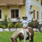 Ein Kind mit Hut sitzt barfuß auf einem kleinen Pony, das auf einer Wiese mit einem gelben Haus und Blumen im Hintergrund weidet.