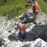 Eine Gruppe von Personen mit Helmen und Kletterausrüstung erklimmt im Freien einen felsigen Abhang, umgeben von Gras und Vegetation.