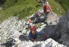 Eine Gruppe von Personen mit Helmen und Kletterausrüstung erklimmt im Freien einen felsigen Abhang, umgeben von Gras und Vegetation.