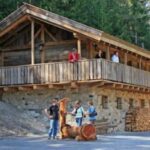 Ein rustikales Holzhaus mit Steinsockel ist von Bäumen umgeben; mehrere Menschen stehen auf dem Balkon und bei den gestapelten Holzscheiten im Freien.