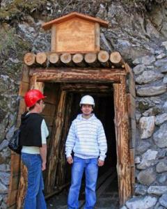 Zwei Personen mit Schutzhelmen stehen am Eingang eines in einen felsigen Abhang gebauten Minenschachts mit Holzrahmen.