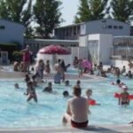 Kinder und Erwachsene schwimmen und spielen in einem überfüllten Freibad an einem sonnigen Tag, mit Gebäuden und Bäumen im Hintergrund.