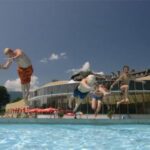 Vier Kinder springen an einem sonnigen Tag in ein Freibad, während im Hintergrund Gebäude und eine Kirche zu sehen sind.