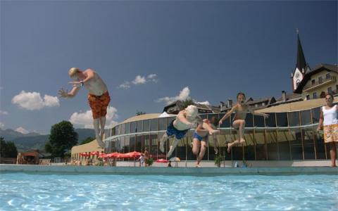 Vier Kinder springen an einem sonnigen Tag in ein Freibad, während im Hintergrund Gebäude und eine Kirche zu sehen sind.