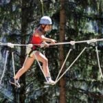 Ein Kind, das einen Helm und einen Klettergurt trägt, durchquert einen Hochseilgarten im Freien, umgeben von hohen Bäumen.