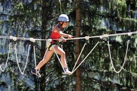 Ein Kind, das einen Helm und einen Klettergurt trägt, durchquert einen Hochseilgarten im Freien, umgeben von hohen Bäumen.