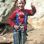 Ein kleines Kind, das einen Helm und ein Gurtzeug trägt, zeigt den Daumen nach oben, während es im Freien in der Nähe einer felsigen Oberfläche steht.