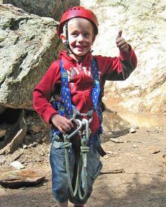 Ein kleines Kind, das einen Helm und ein Gurtzeug trägt, zeigt den Daumen nach oben, während es im Freien in der Nähe einer felsigen Oberfläche steht.