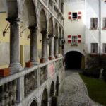 Steinbögen und Säulen säumen einen Balkon mit Blick auf einen Hof mit Kopfsteinpflaster und ein weißes Gebäude mit roten und weißen Fensterläden.