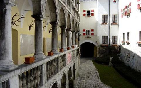 Steinbögen und Säulen säumen einen Balkon mit Blick auf einen Hof mit Kopfsteinpflaster und ein weißes Gebäude mit roten und weißen Fensterläden.