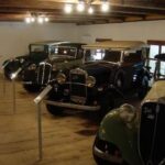 Vier Oldtimer sind in einem Innenraum auf einem Holzboden unter Deckenbeleuchtung ausgestellt, jeder hinter einem Informationsständer aus Metall.