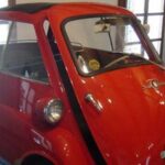 Ein roter BMW Isetta Kleinwagen mit einer einzigen Vordertür ist in der Nähe von Fenstern geparkt.
