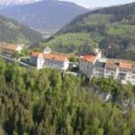 Ein großer historischer Gebäudekomplex mit roten Dächern liegt an einem Hang, umgeben von grünen Wäldern und Bergen.