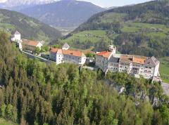 Ein großer historischer Gebäudekomplex mit roten Dächern liegt an einem Hang, umgeben von grünen Wäldern und Bergen.