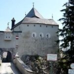 Ein steinernes Schloss mit schrägem Dach, runden Fenstern und Fahnen steht am Ende einer Brücke, umgeben von Bäumen und Schildern am Eingang.