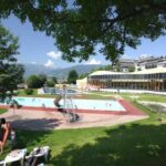 Freibad mit Rutsche, umgeben von Gras, Bäumen und ein paar Menschen; im Hintergrund sind Gebäude und Berge zu sehen.
