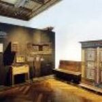 Ausstellungsraum mit antiken Holzmöbeln, darunter ein Kleiderschrank, eine Bank und Schränke, vor einer dunklen Wand mit erklärendem Text.