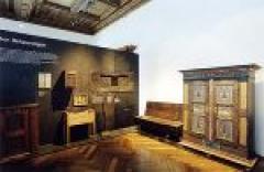 Ausstellungsraum mit antiken Holzmöbeln, darunter ein Kleiderschrank, eine Bank und Schränke, vor einer dunklen Wand mit erklärendem Text.