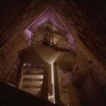 Eine Metalltreppe mit Geländer führt im Inneren einer hohen, dreieckigen Steinstruktur hinauf, die von einer weichen, indirekten Beleuchtung erhellt wird.