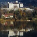 Ein großes, weißes Gebäude steht auf einem von Bäumen umgebenen Hügel, dessen Spiegelbild sich im ruhigen Wasser spiegelt.