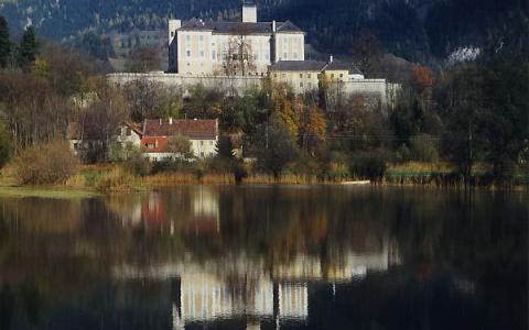 Ein großes, weißes Gebäude steht auf einem von Bäumen umgebenen Hügel, dessen Spiegelbild sich im ruhigen Wasser spiegelt.