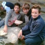 Drei Jungen sitzen im Freien auf großen Felsen, halten und untersuchen Steinplatten. Andere Kinder sind im Hintergrund zu sehen.