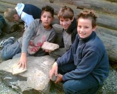 Drei Jungen sitzen im Freien auf großen Felsen, halten und untersuchen Steinplatten. Andere Kinder sind im Hintergrund zu sehen.