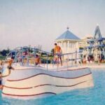 Die Menschen schwimmen und spielen um eine wellenförmige, weiße Kletterstruktur in einem Wasserpark im Freien mit Gebäuden im Hintergrund.