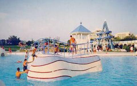 Die Menschen schwimmen und spielen um eine wellenförmige, weiße Kletterstruktur in einem Wasserpark im Freien mit Gebäuden im Hintergrund.