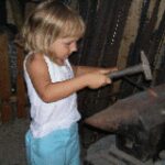 Ein junges Kind in einem weißen Tank-Top und blauen Shorts benutzt einen Hammer auf einem Amboss in einer Werkstattumgebung.