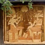 Holzschnitzerei, die eine altägyptische Szene mit zwei sitzenden Figuren, einem Kind, Hieroglyphen und einer Sonne mit Strahlen darstellt, montiert an einer Außenwand.