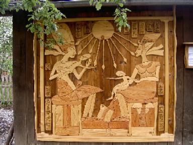 Holzschnitzerei, die eine altägyptische Szene mit zwei sitzenden Figuren, einem Kind, Hieroglyphen und einer Sonne mit Strahlen darstellt, montiert an einer Außenwand.