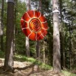 Eine große, rote Spiralinstallation hängt zwischen Bäumen in einem Wald, durch deren Blätter das Sonnenlicht fällt.