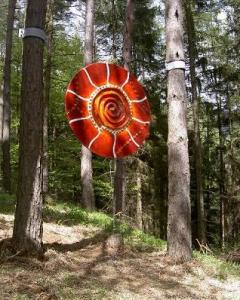 Eine große, rote Spiralinstallation hängt zwischen Bäumen in einem Wald, durch deren Blätter das Sonnenlicht fällt.