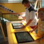 Zwei Personen verwenden Touchscreen-Displays an einem Holztisch in einem hell erleuchteten Innenraum.