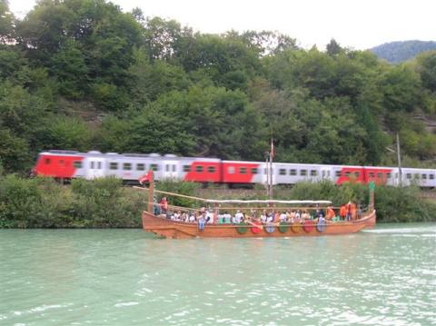Ein Holzboot mit Menschen an Bord schwimmt auf einem Fluss, während im Hintergrund ein rot-weißer Zug auf Gleisen vorbeifährt, umgeben von Grünzeug.