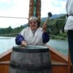 Eine Person mit gehörntem Helm schlägt mit einem Schlegel eine Fasstrommel auf einem Holzboot an, im Hintergrund sind Wasser und Bäume zu sehen.