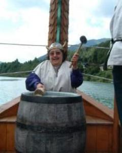 Eine Person mit gehörntem Helm schlägt mit einem Schlegel eine Fasstrommel auf einem Holzboot an, im Hintergrund sind Wasser und Bäume zu sehen.