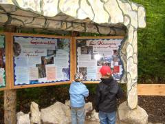 Zwei Kinder stehen vor den Informationstafeln im Freien und lesen die Schilder unter einem Unterstand aus Stein und Holz.