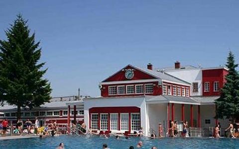 Menschen schwimmen in einem Freibad vor einem rot-weißen Gebäude mit großen Fenstern und einer Uhr an der oberen Fassade, unter einem klaren blauen Himmel.