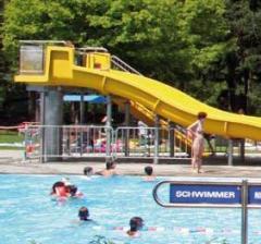 Menschen schwimmen in einem Freibad in der Nähe einer gelben Wasserrutsche; eine Frau steht am Eingang der Rutsche. Im Hintergrund sind Bäume und ein "Schwimmer"-Schild zu sehen.