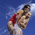 Zwei Personen in Badeanzügen lachen und planschen im Wasser vor einem strahlend blauen Himmel.