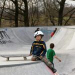 Ein kleines Kind mit einem Helm sitzt mit einem Skateboard am Rand einer Skateparkrampe, während ein anderes Kind in einem grünen T-Shirt in der Nähe steht.
